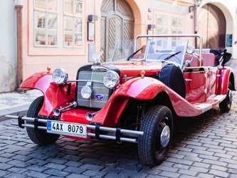 en:A bright red vintage convertible is parked on a cobblestone street in front of historic buildings in Prague. ;fr:Une voiture historique rouge vif à ciel ouvert est garée sur une rue pavée devant des bâtiments historiques de Prague.