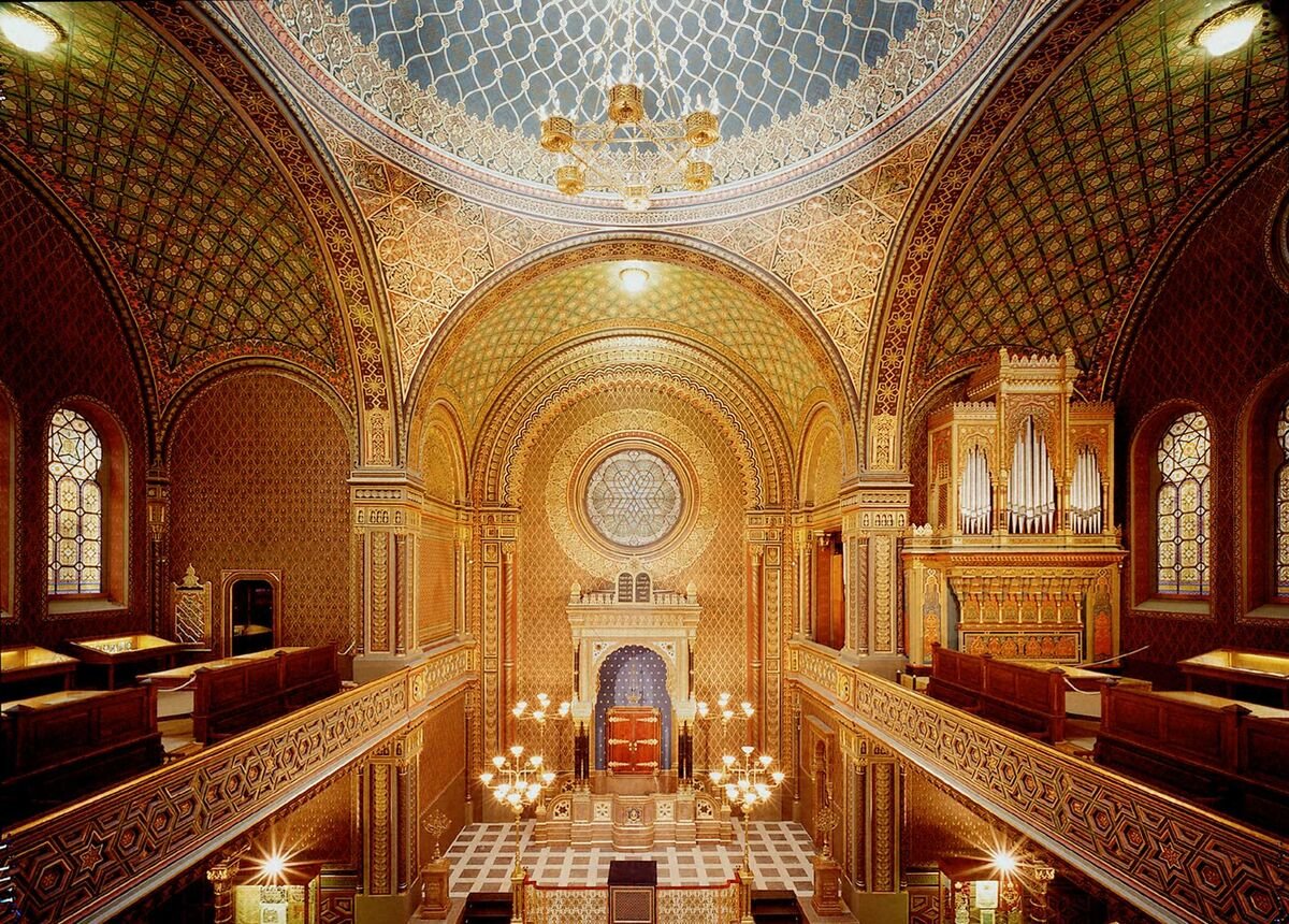 A majestic hall with walls carved in gold featuring Hispano-Moorish motifs, and circular stained-glass windows with designs