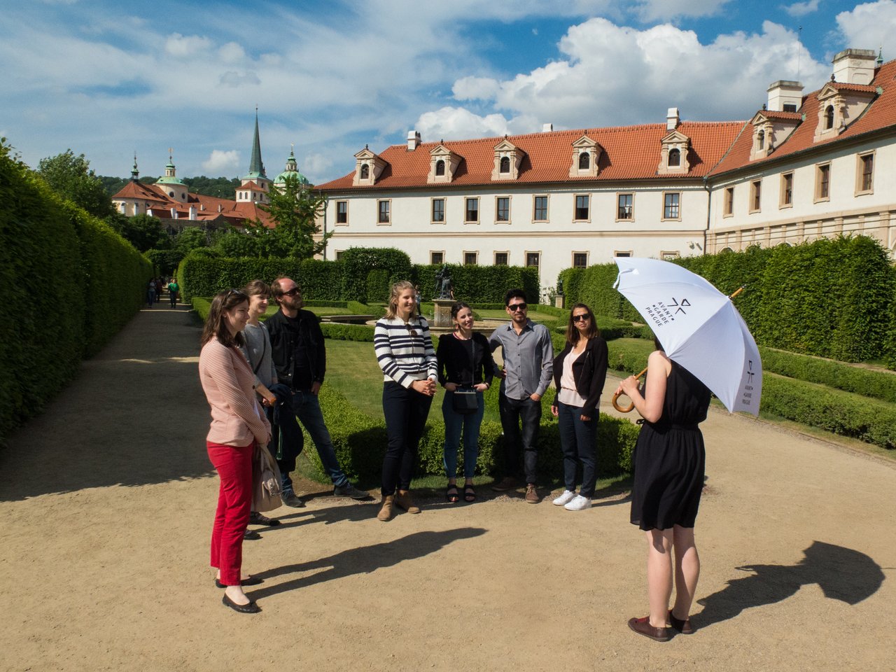 A tour guide, holding an umbrella, is speaking to seven people in a beautiful garden