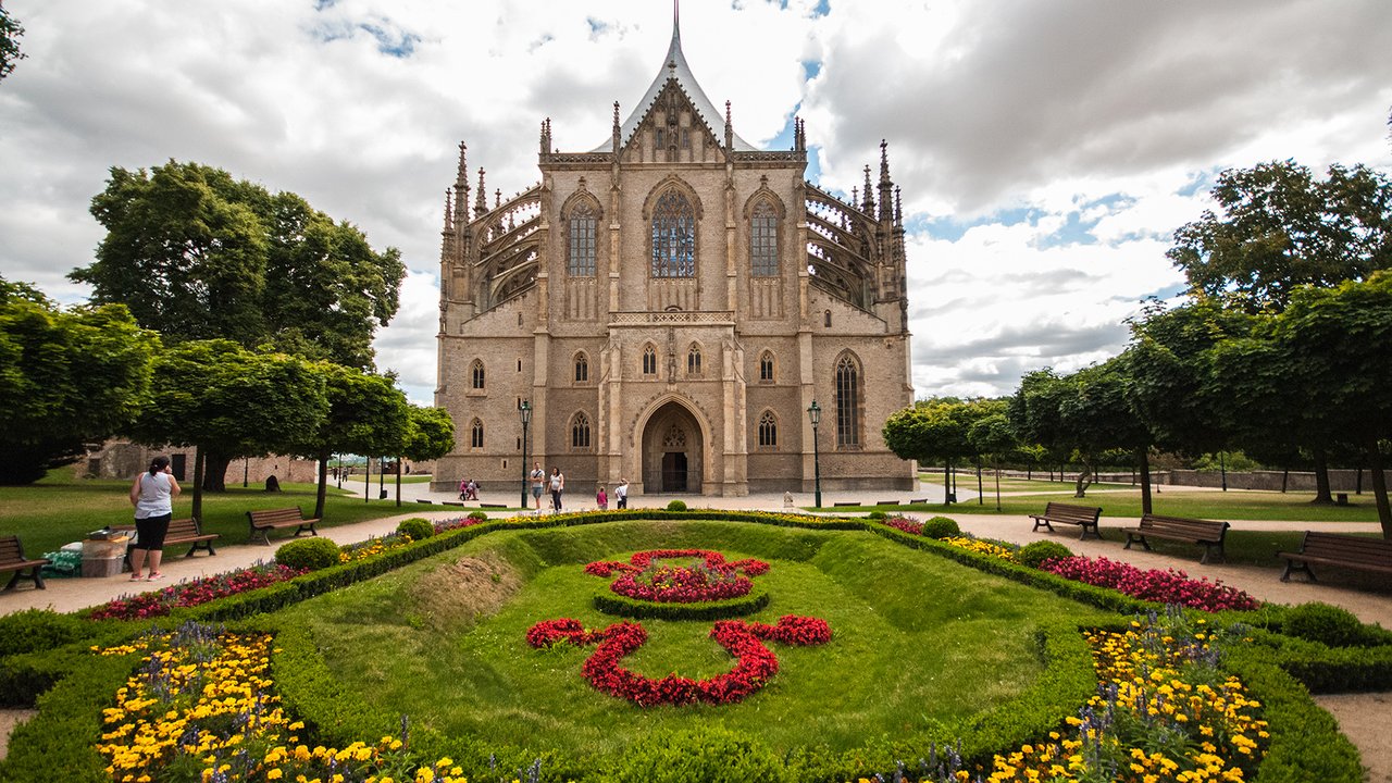 A large stone church with very pointed roofs rises behind a garden of green lawn and colorful flowers.