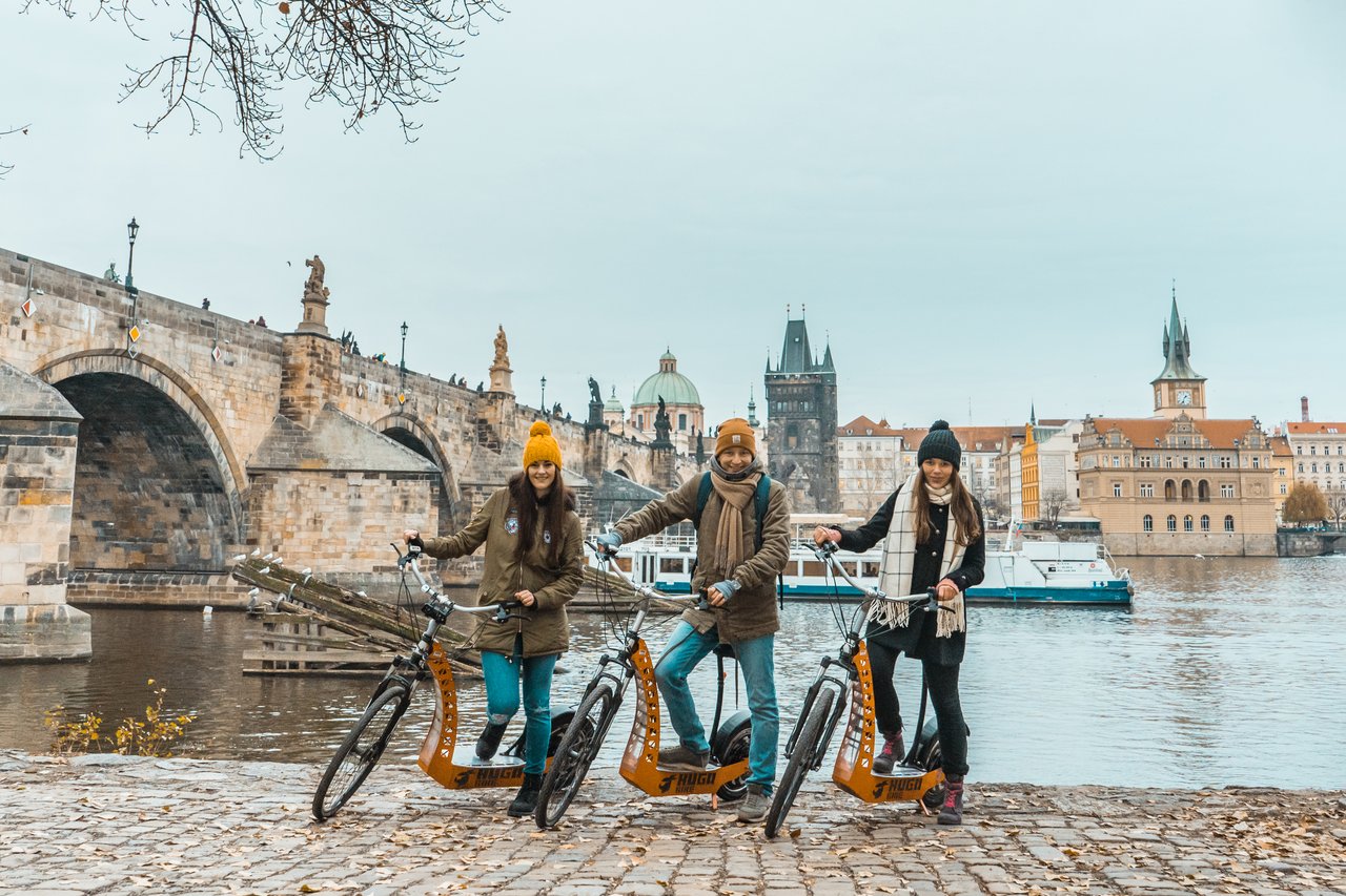 3 people on electric scooters in the fall,with the river flowing beneath an iconic old bridge in the background