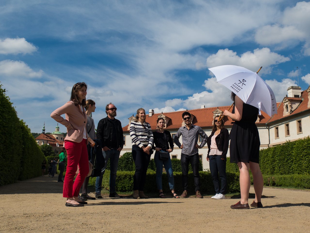 A tour guide, umbrella in hand, is speaking to seven people in a beautiful garden under a sunny sky.