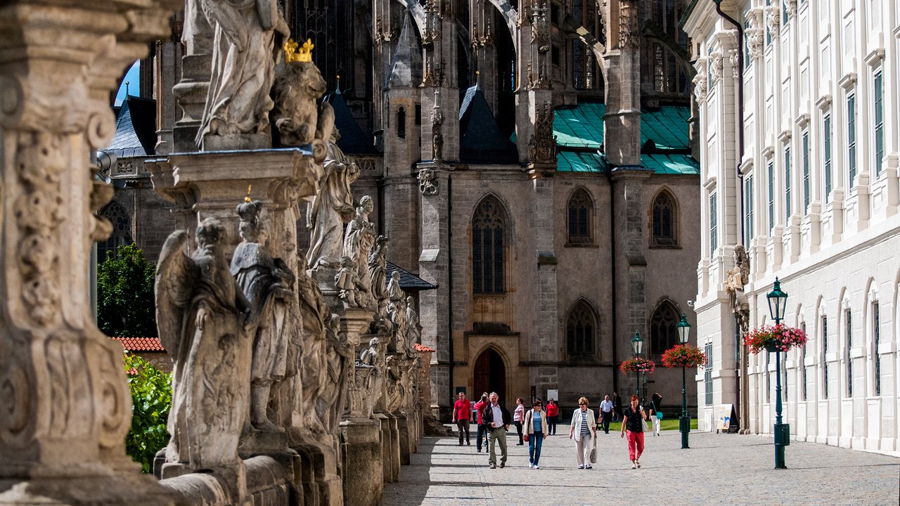 A sunny avenue is lined with a row of dark stone statues, with a large church in the background