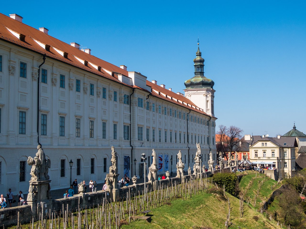A long white building with an orange tiled roof stretches beside a low wall adorned with stone statues, overlooking a verdant hill.
