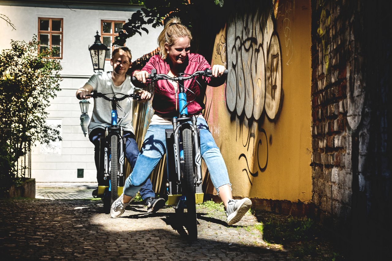 Two smiling people on an electric scooter riding down a small street lined with stone walls