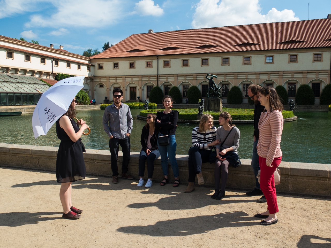 A tour guide, holding an umbrella, is speaking to seven people sitting in front of a fountain under a sunny sky.