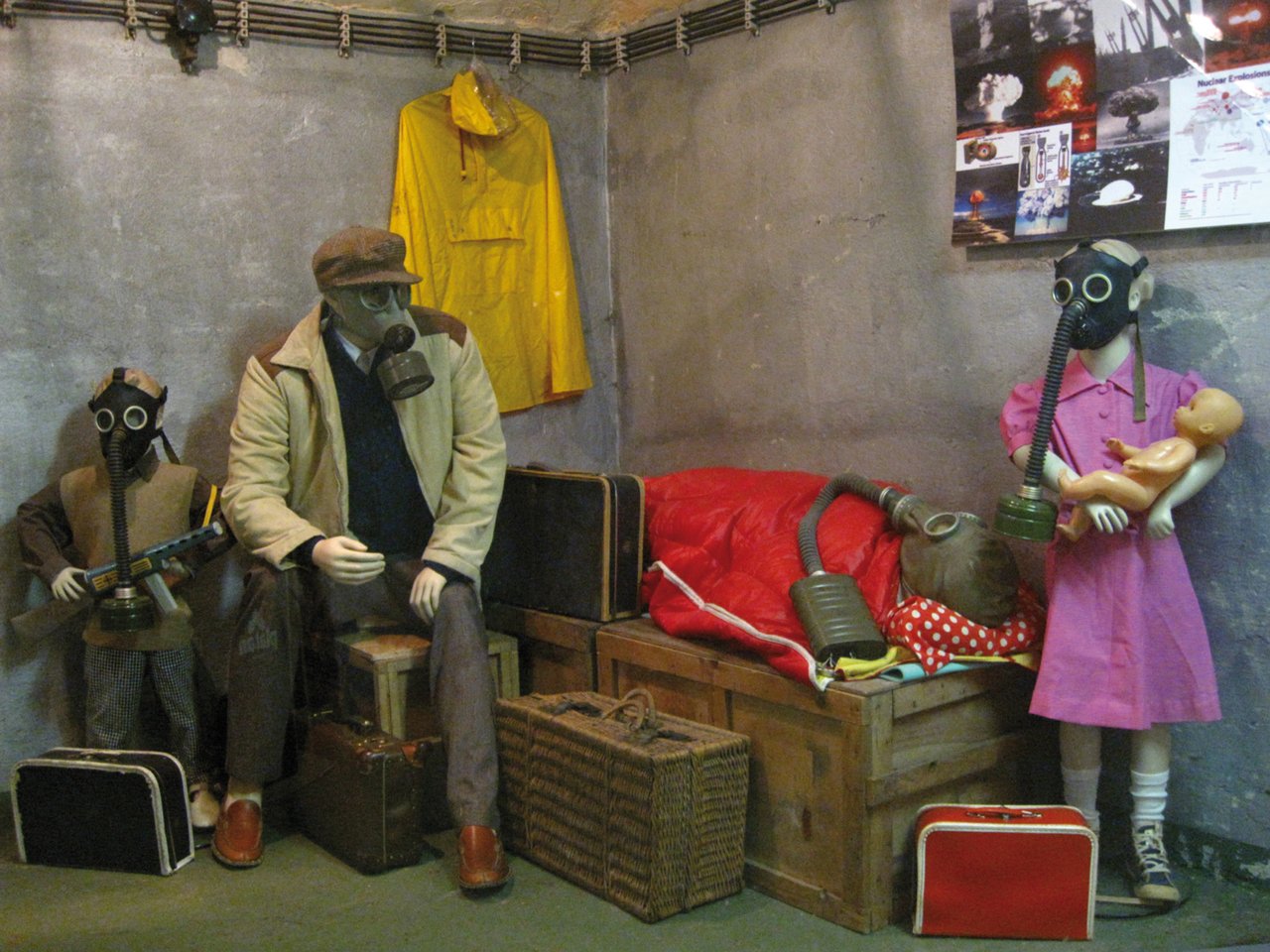 A family wearing gas masks in a concrete room where a poster depicting bombs is hanging
